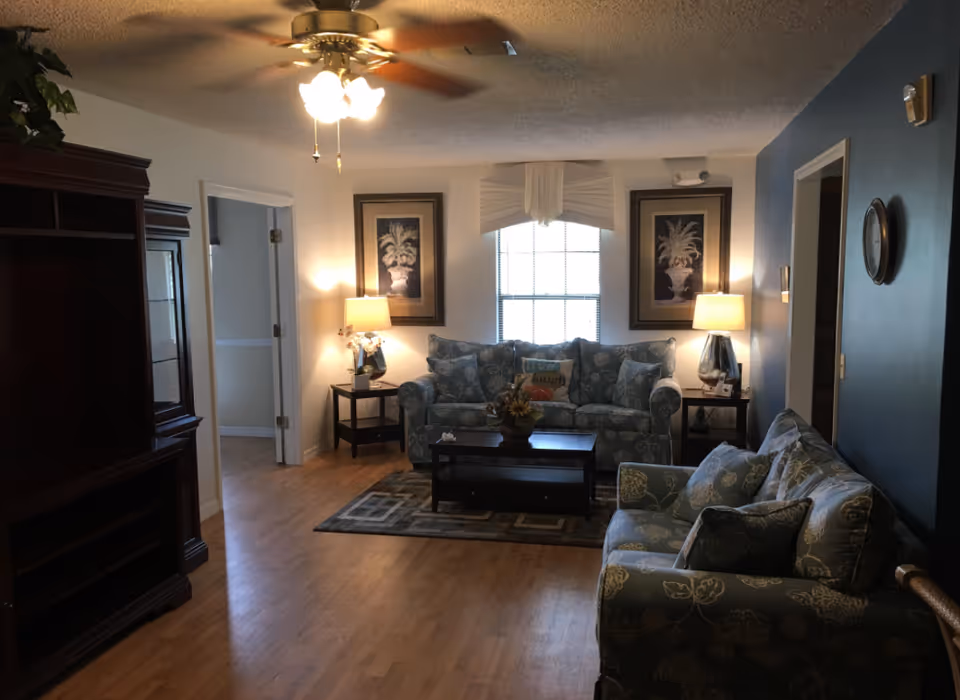 A cozy living room with two blue patterned sofas, a wooden coffee table, two side tables with lamps, framed artwork on the walls, a ceiling fan with lights, and a window with a decorative valance letting in natural light.