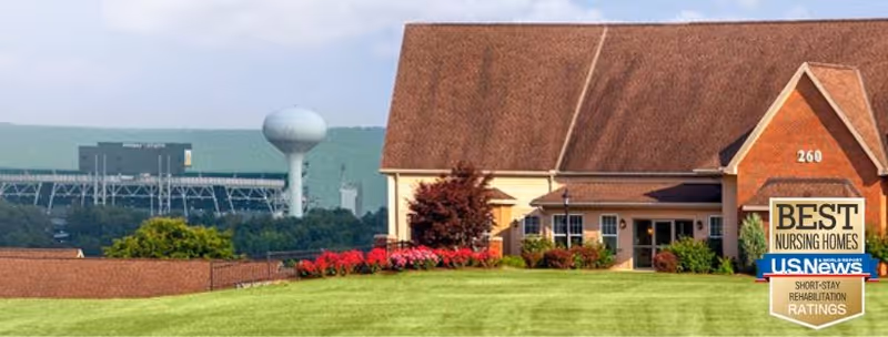 Front exterior of The Village At Penn State showing a lawn and flowerbeds in front of a brick-faced building, with a water tower and stadium visible in the background.