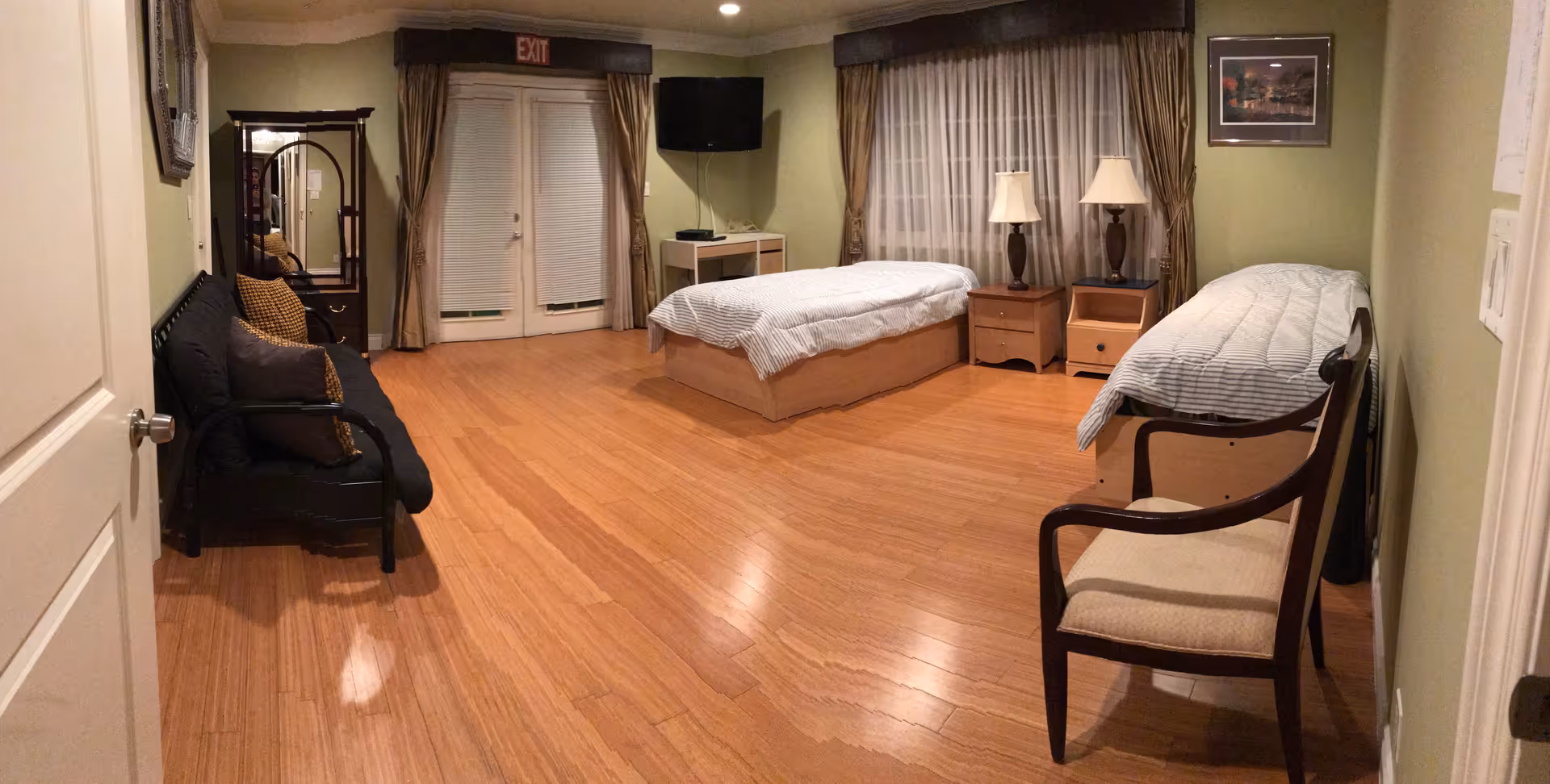 A spacious bedroom with two single beds covered in white and gray striped bedding. Between the beds is a wooden nightstand with two lamps. A wooden chair with a cushioned seat is placed near the bed on the right. On the left side of the room, there is a black futon sofa with several pillows, and a tall mirror with a wooden frame. The room has light green walls, wooden flooring, and large windows with sheer white curtains and brown drapes. A flat-screen TV is mounted on the wall above a small white table. Double doors with blinds and an exit sign above them are visible at the far end of the room.
