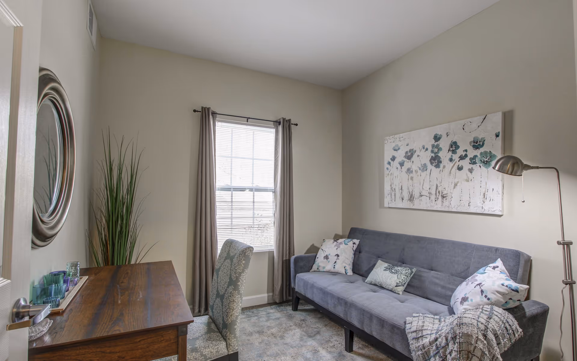 Small living room with a gray sofa and throw pillows, a wooden desk and chair, floor lamp, wall art, and a window with curtains.