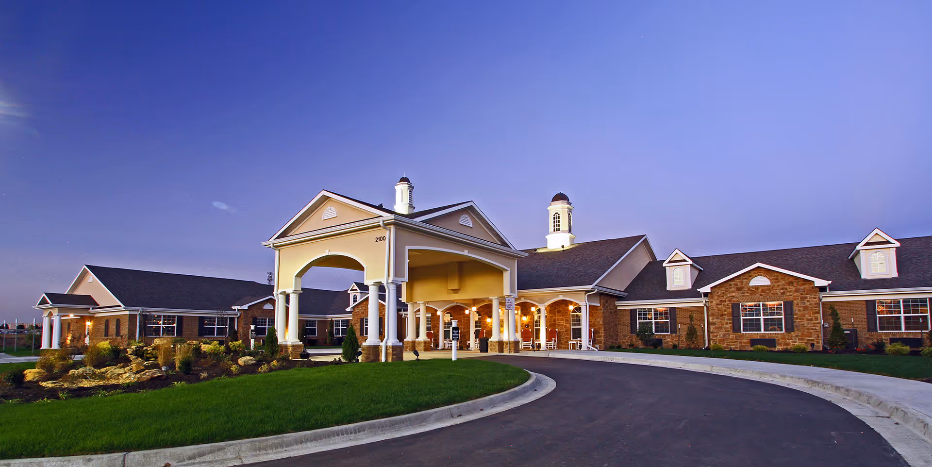 Exterior view of Benton House of Raymore senior living facility at dusk, showing a large building with brick and stone facade, white columns, and a covered entrance with a circular driveway and landscaped greenery.