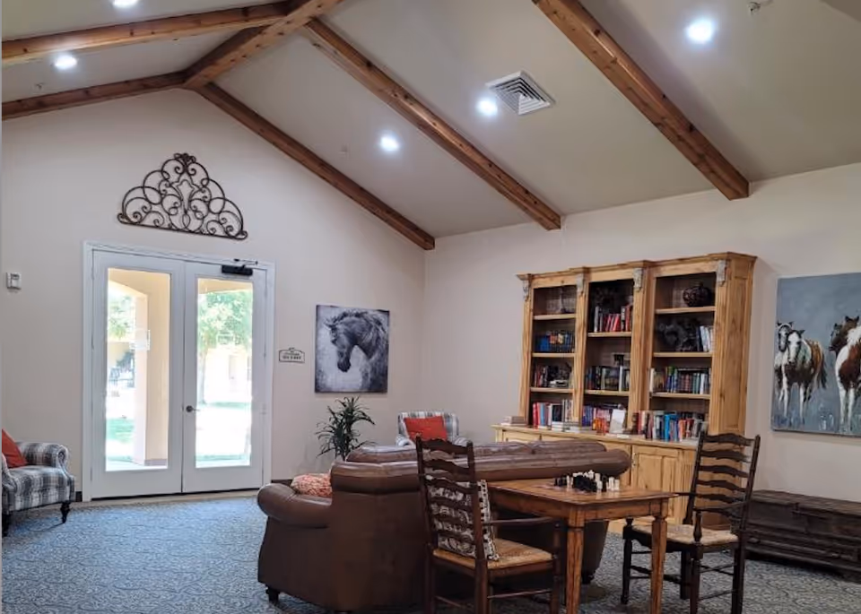 A cozy senior living common area with a high ceiling featuring wooden beams. The room has a brown leather sofa, two wooden chairs, a wooden bookshelf filled with books, and artwork of horses on the walls. There is a double glass door leading outside, and a patterned carpet covers the floor.