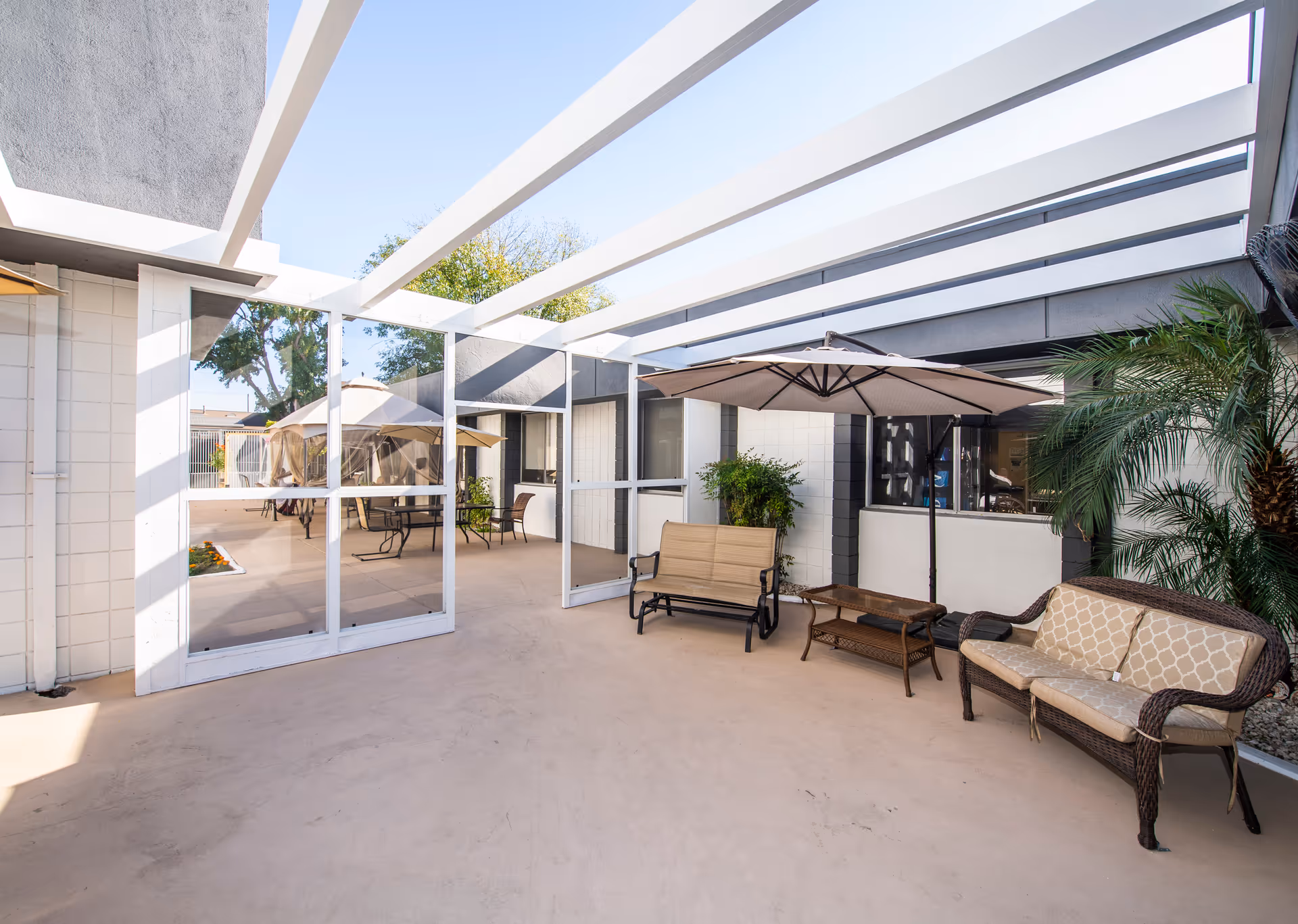 Outdoor patio area at Sierra View Care Center with seating including cushioned wicker chairs, a bench, a table with an umbrella, potted plants, and a pergola overhead providing partial shade.