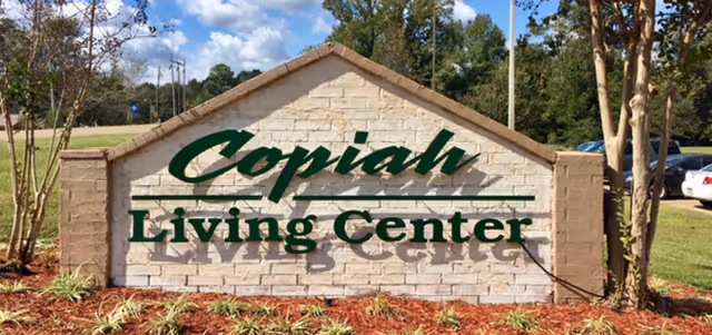 A large brick sign with a triangular top displaying the name 'Copiah Living Center' in green letters, surrounded by landscaped plants and trees with a parking lot and cars visible in the background under a partly cloudy sky.