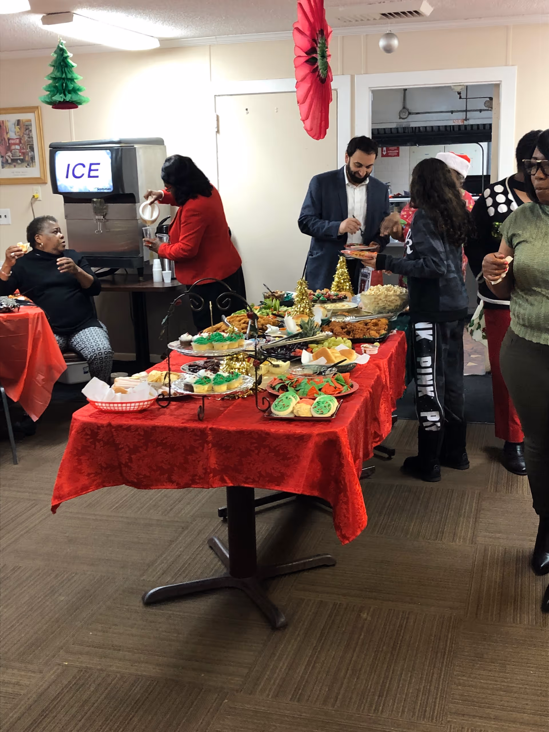 A festive indoor gathering at an assisted living facility with a table covered in a red tablecloth displaying various holiday-themed snacks and treats. Several people are serving themselves food and drinks, with holiday decorations hanging from the ceiling and a large ice dispenser in the background.