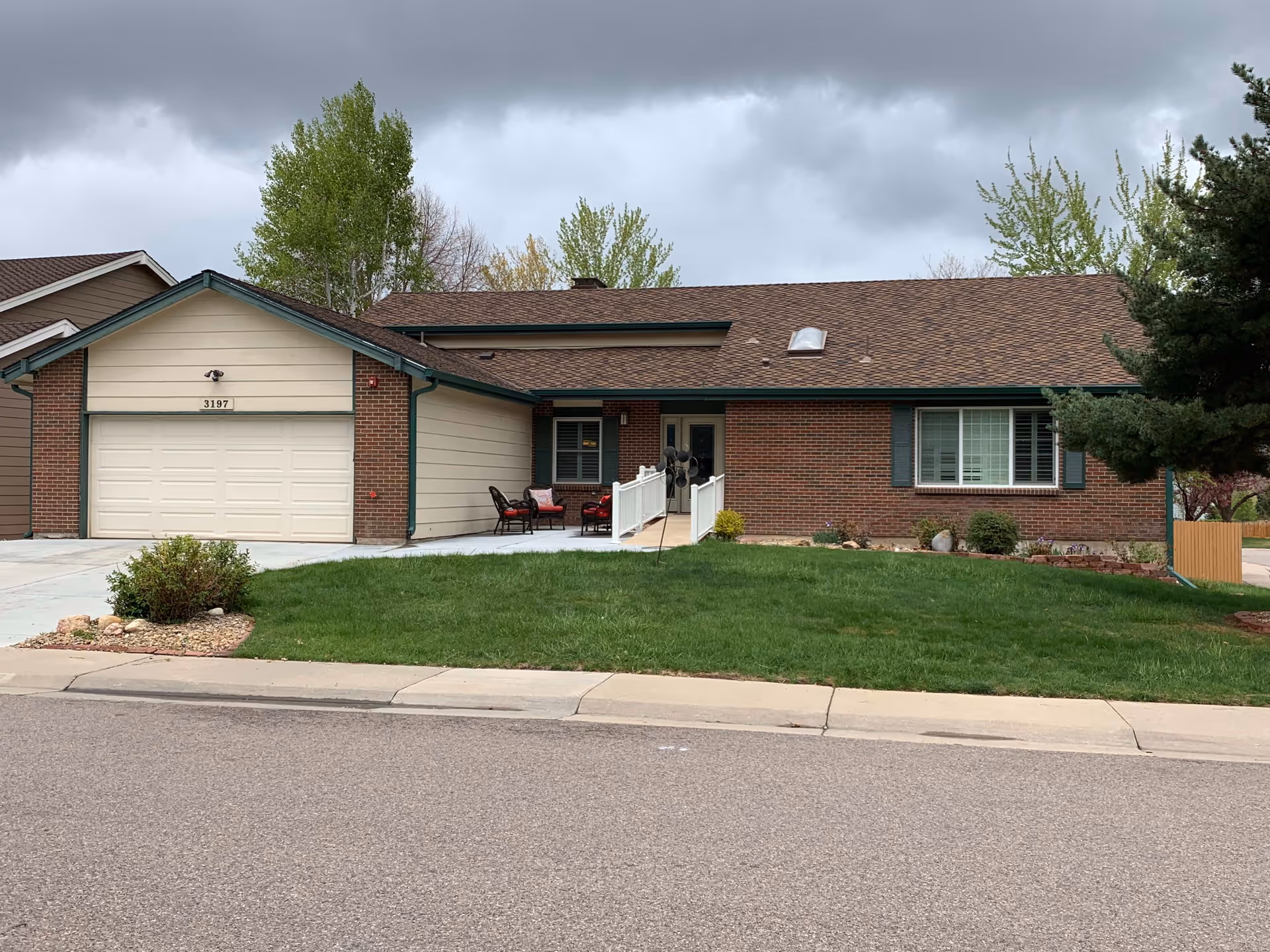 Single-story brick and siding house with a two-car garage, a small front porch with outdoor chairs, a green lawn, and trees in the background under a cloudy sky.