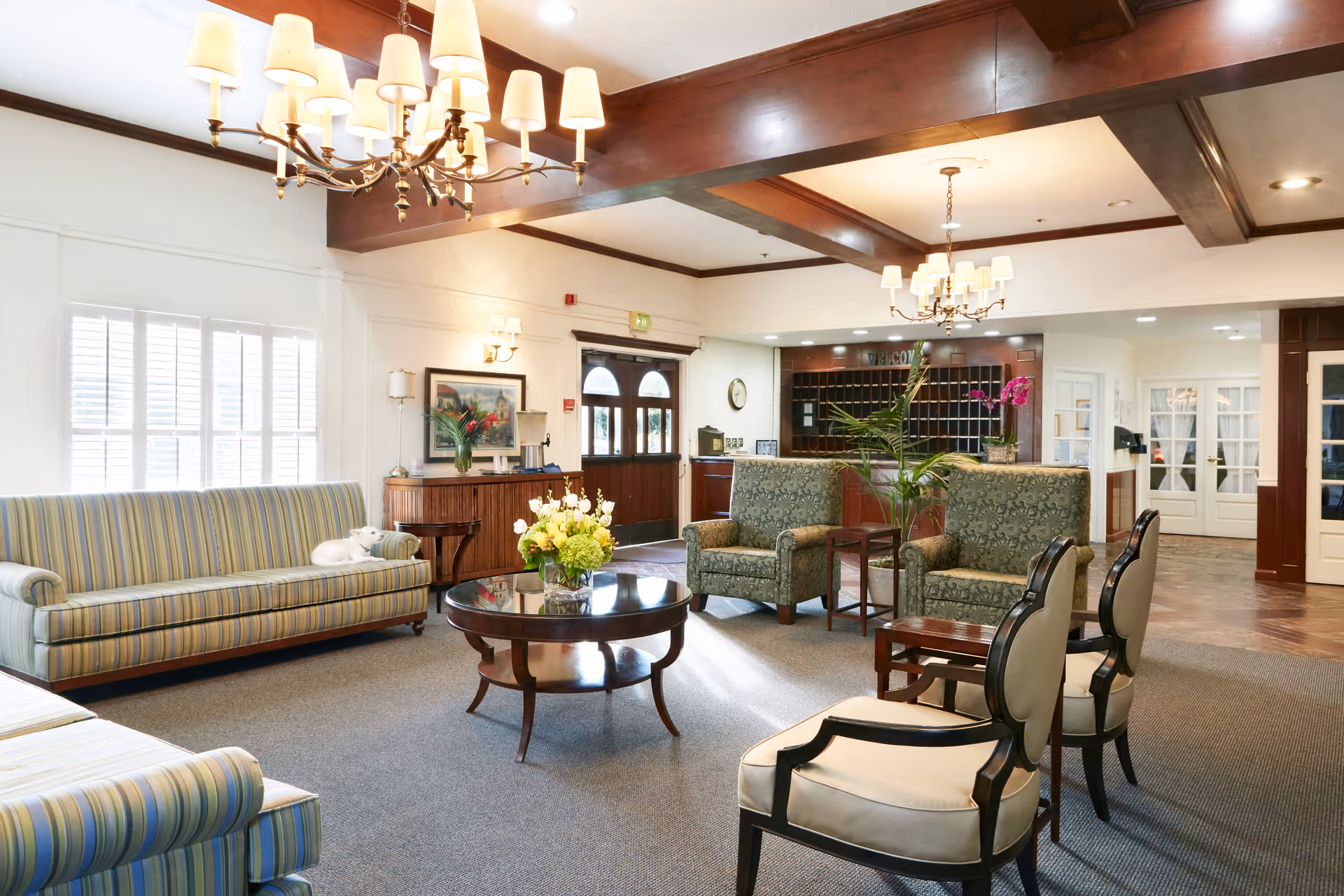 A bright and spacious living room area in a senior living facility with striped sofas, patterned armchairs, and cream-colored chairs arranged around a round wooden coffee table with a flower arrangement. The room features wooden ceiling beams, chandeliers, large windows with white shutters, and a reception desk in the background.