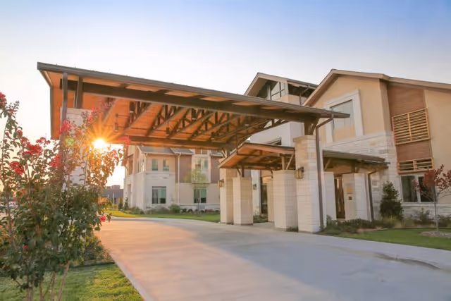 Exterior view of The Healthcare Resort of Plano building entrance with a covered driveway, stone pillars, and landscaped greenery under a clear sky with the sun setting in the background.