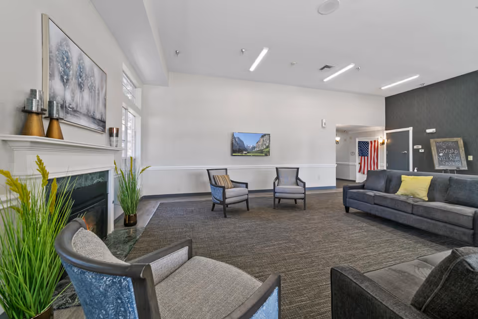 A spacious living room area in a senior living facility with a gray sofa, two gray armchairs, a fireplace with decorative items on the mantel, and a flat-screen TV mounted on the wall. There are large windows letting in natural light, a carpeted floor, and a chalkboard sign near the entrance that reads 'Cariad at Village Creek'.