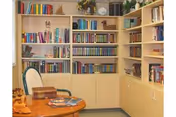 A cozy library or reading room with light wood bookshelves filled with books and decorative items. There is a round wooden table with a few books and a chair with a green frame and white cushion. The room has a warm and inviting atmosphere.