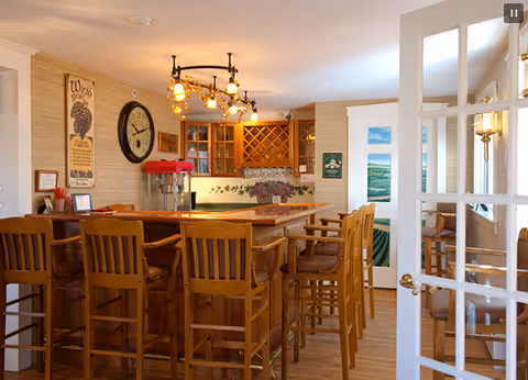 Interior view of a cozy bar area with wooden high chairs lined up along a wooden counter. The back wall features a wine rack, cabinets, a large clock, and a popcorn machine. Warm lighting fixtures hang from the ceiling, and a glass-paned door is partially open on the right side.
