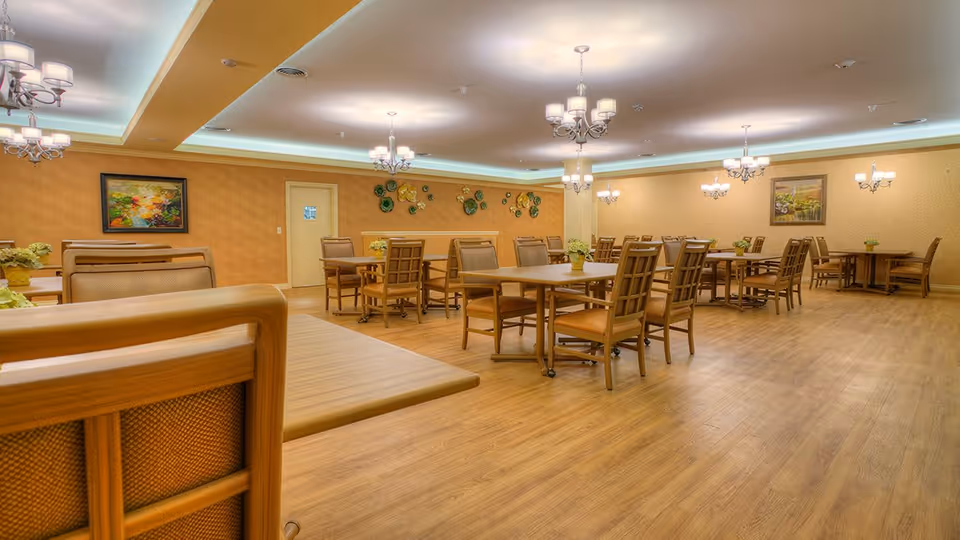 A spacious dining room with multiple wooden tables and chairs arranged neatly on a wood floor. The walls are decorated with framed paintings and ornamental wall art. Several chandeliers hang from the ceiling, providing warm lighting to the room.