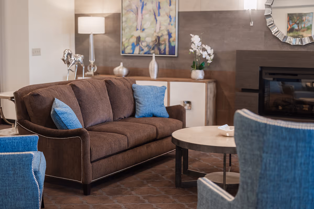 A cozy living room with a brown sofa adorned with blue pillows, a round coffee table, blue upholstered chairs, a sideboard with decorative vases and a white orchid, a floor lamp, a colorful abstract painting on the wall, and a fireplace with a decorative mirror above it.