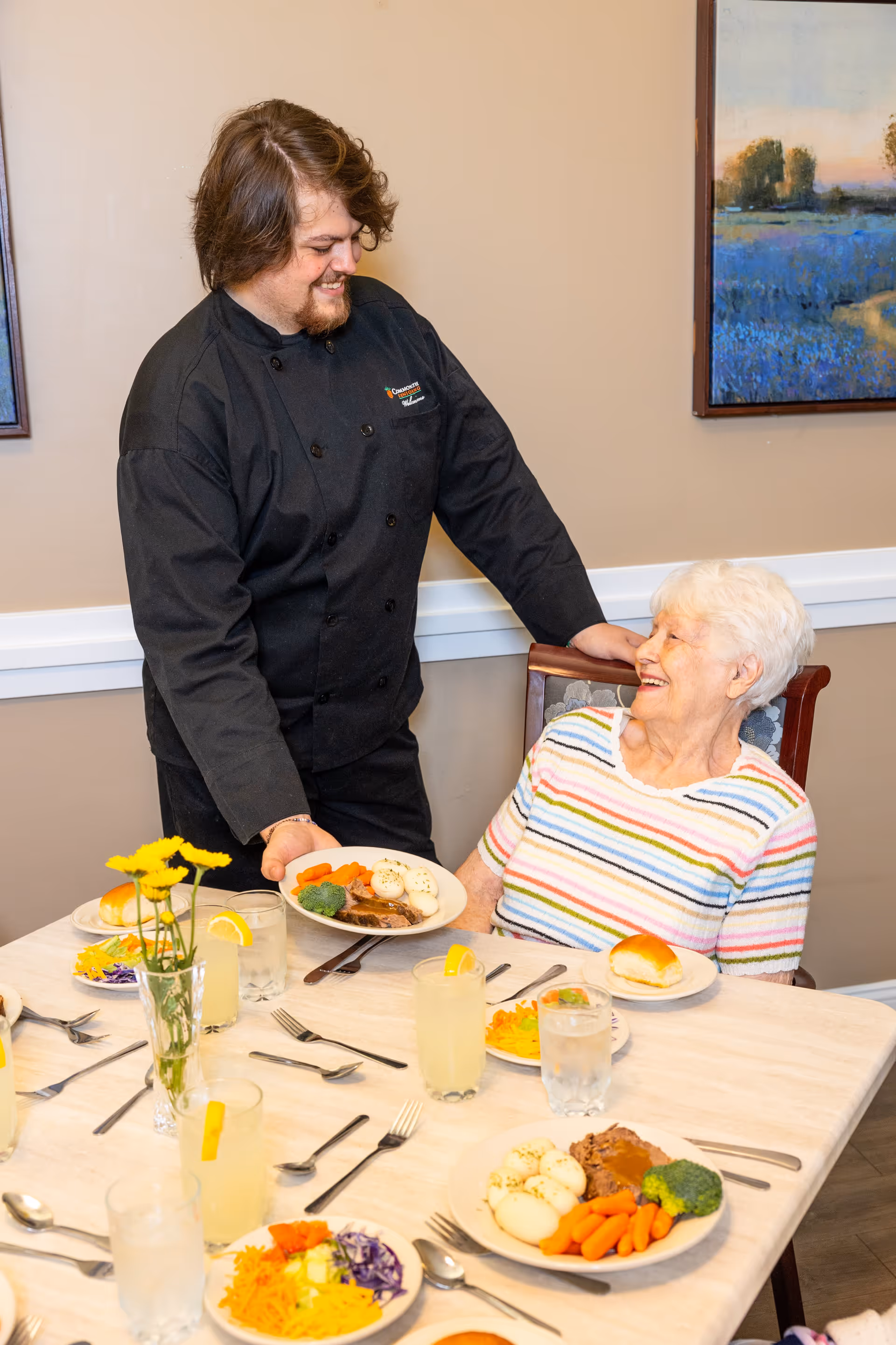 A smiling chef in a black uniform serving a plate of food with meat, carrots, broccoli, and boiled eggs to an elderly woman wearing a striped shirt seated at a dining table set with plates of food, glasses of water and lemonade, and a vase with yellow flowers in a dining room.