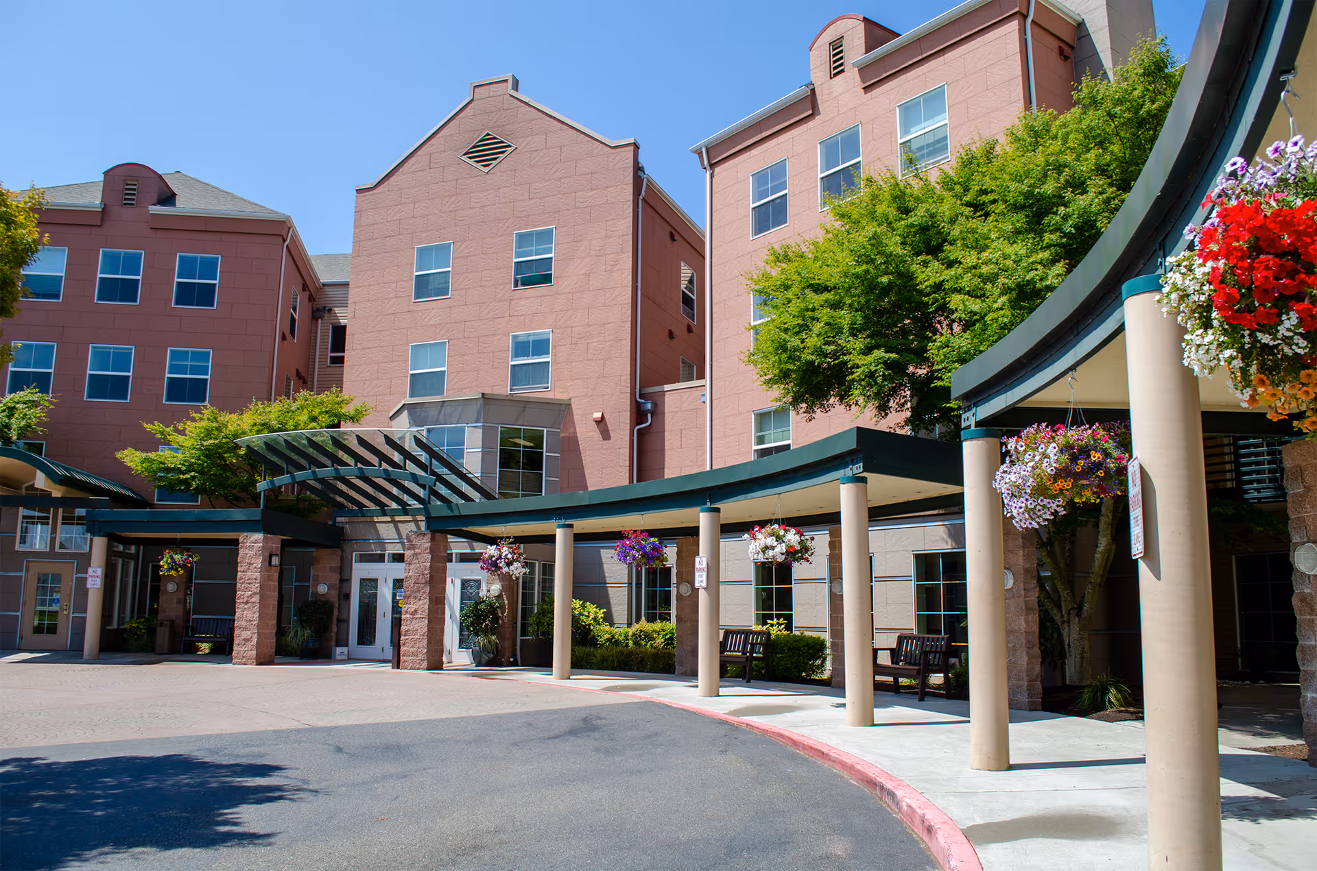 Front entrance of a multi-story brick senior living building with a covered porte-cochere, hanging flower baskets, and benches.
