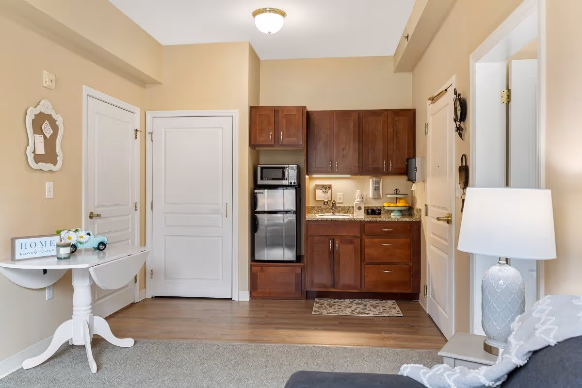 A small kitchenette area with dark wooden cabinets, a mini refrigerator, microwave, and a sink. To the left, there is a white drop-leaf table with a decorative sign that says 'HOME sweet home', a small vase with flowers, and a candle. The walls are painted beige, and there are two white doors visible. On the right side, part of a gray couch with a white and gray throw blanket and a white table lamp on a side table are visible.