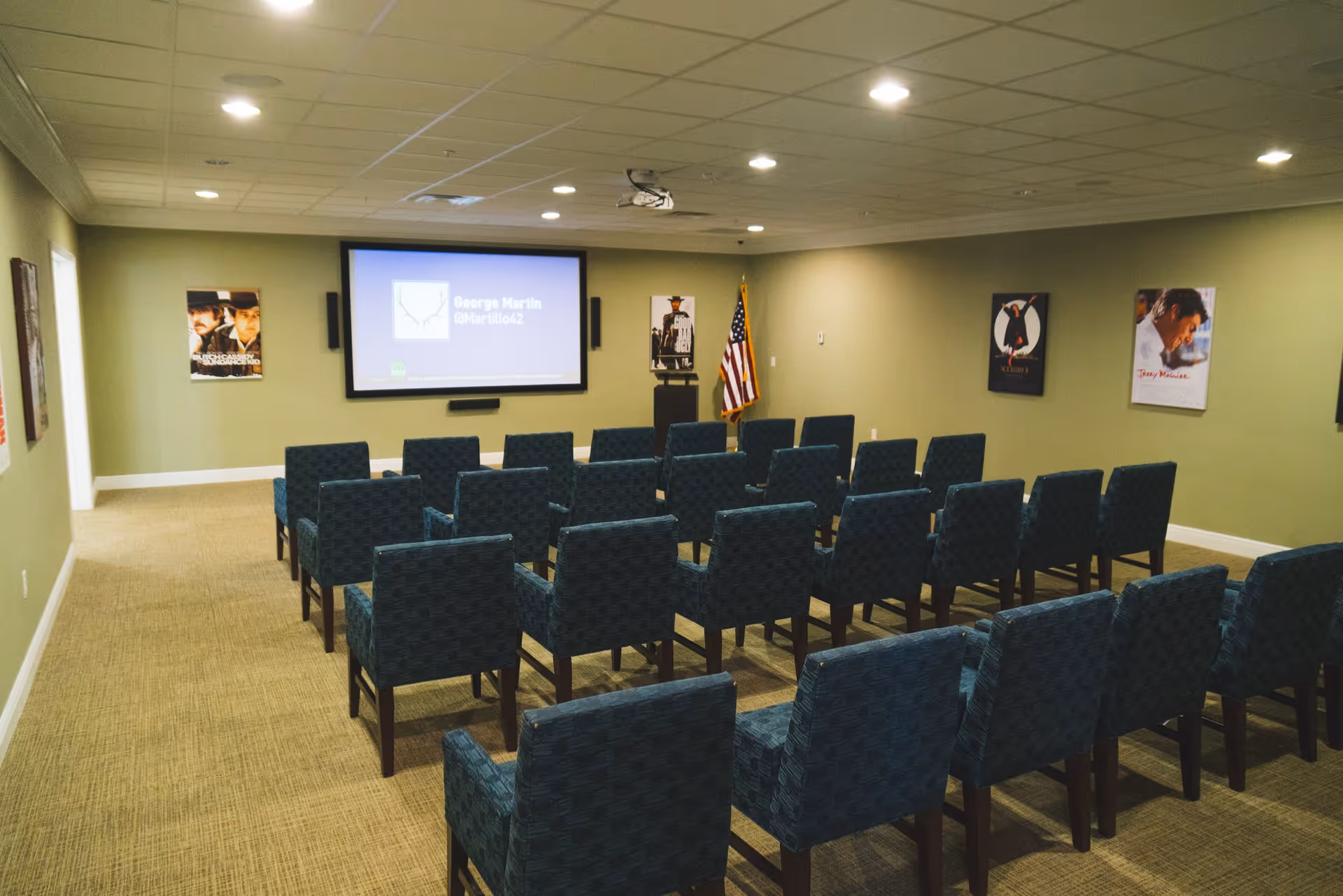 A small theater or presentation room with rows of blue upholstered chairs facing a large screen on the front wall. The walls are light green and decorated with movie posters. An American flag stands near the screen, and the ceiling has recessed lighting.