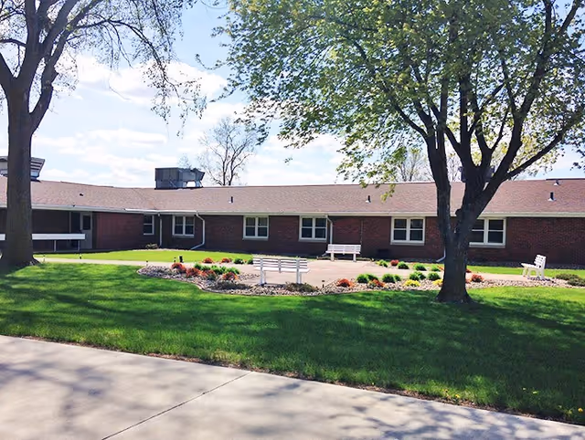 Outdoor view of a single-story brick building with several windows, surrounded by green grass, trees, and a landscaped area with benches and small bushes under a partly cloudy sky.