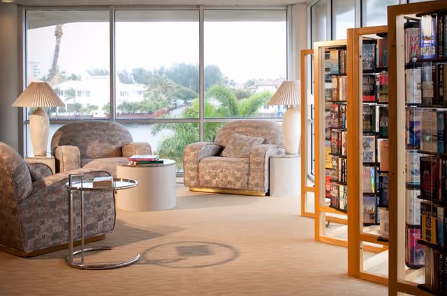 Sunlit lounge with upholstered armchairs, side tables, lamps, and bookshelves in front of large windows overlooking water and palm trees.