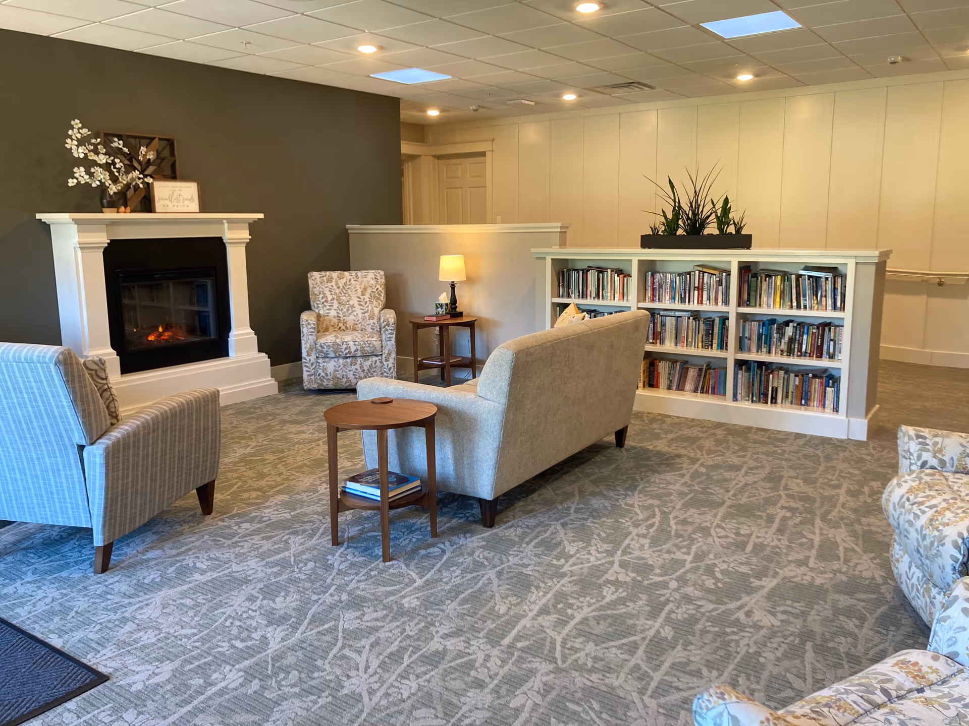 A cozy living room area in a retirement community featuring a lit fireplace with a white mantel, three upholstered armchairs, a beige sofa, a small wooden side table with books, a lamp on a side table, and a built-in bookshelf filled with books. The room has a patterned carpet and neutral-colored walls.