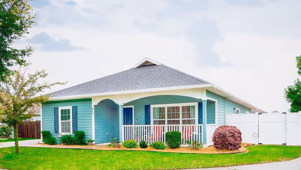 Single-story blue house with white trim and a front porch with two red chairs, surrounded by green grass, bushes, and a small tree under a partly cloudy sky.
