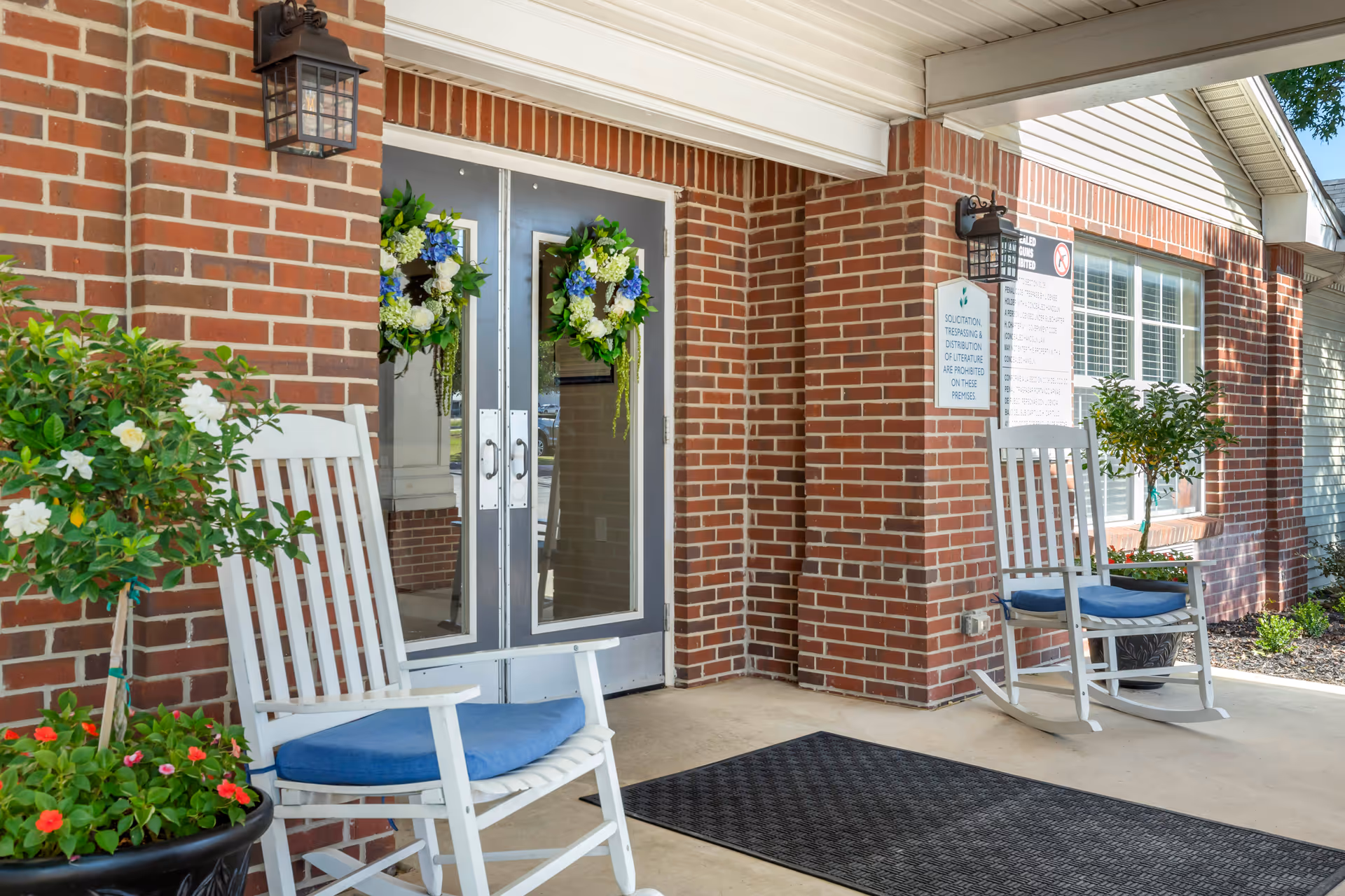 Entrance to a brick building with double glass doors decorated with floral wreaths. Two white rocking chairs with blue cushions are placed on either side of the entrance, along with potted plants. A black outdoor mat lies in front of the doors, and there are wall-mounted lantern-style lights above the chairs.