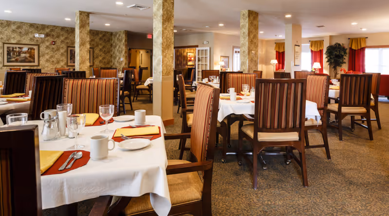 A spacious dining room with multiple tables covered in white tablecloths, set with plates, cups, glasses, and silverware. The chairs have striped upholstery and wooden frames. The room has beige walls with decorative columns, carpeted floors, and windows with red and yellow curtains. Lamps and framed artwork decorate the space.