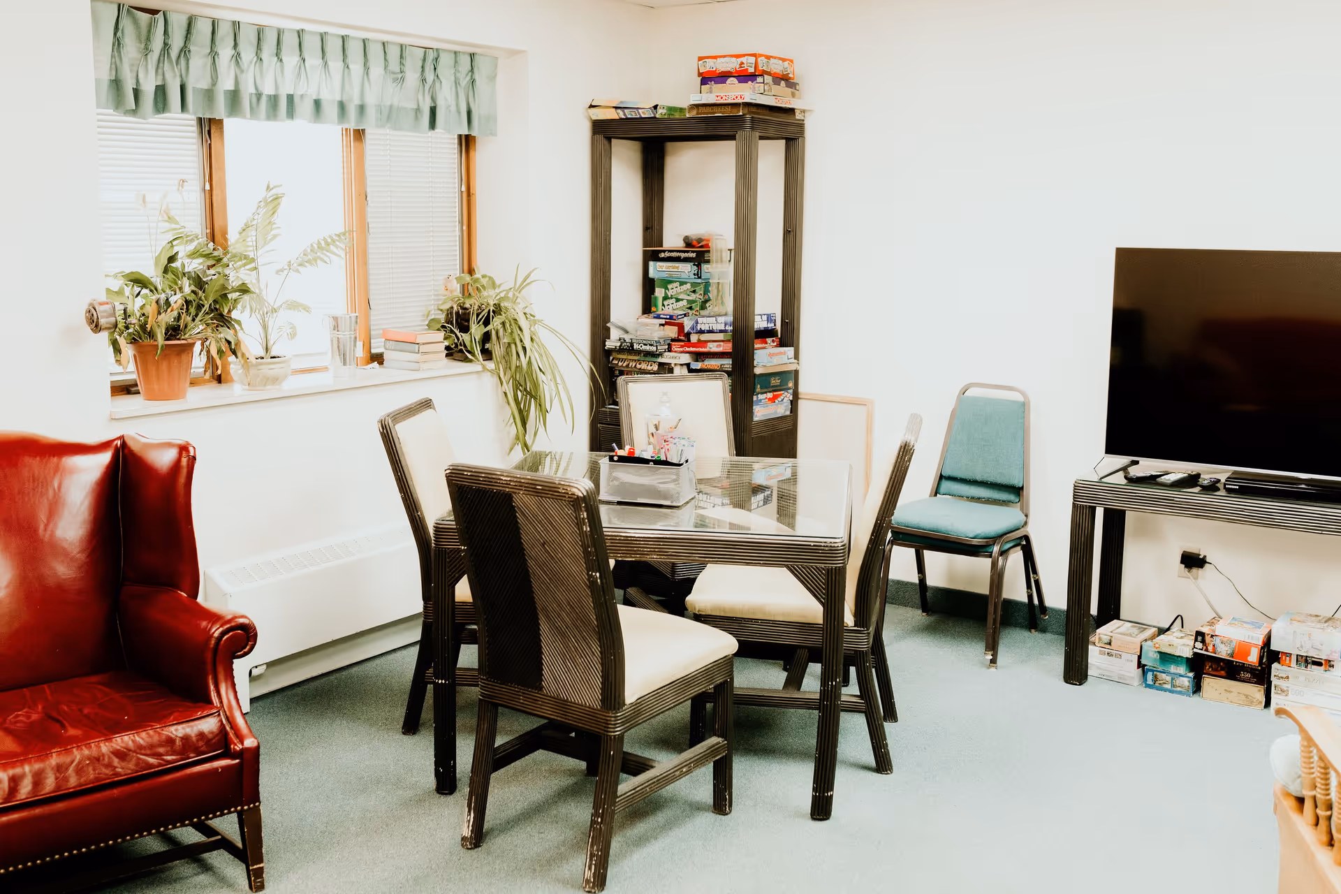 A cozy room with a glass-top table surrounded by four chairs, a red leather armchair, a green cushioned chair, and a black shelving unit filled with board games. There are plants on the windowsill and a large flat-screen TV on a stand with more board games underneath.