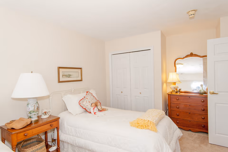 A cozy bedroom with a single bed covered in a white quilt and decorative pillows, including a doll. There is a wooden nightstand with a floral lamp, a mug, and a wooden bowl. A framed picture hangs on the beige wall above the bed. Across from the bed is a wooden dresser with a mirror and a lamp, next to a closed white closet door.