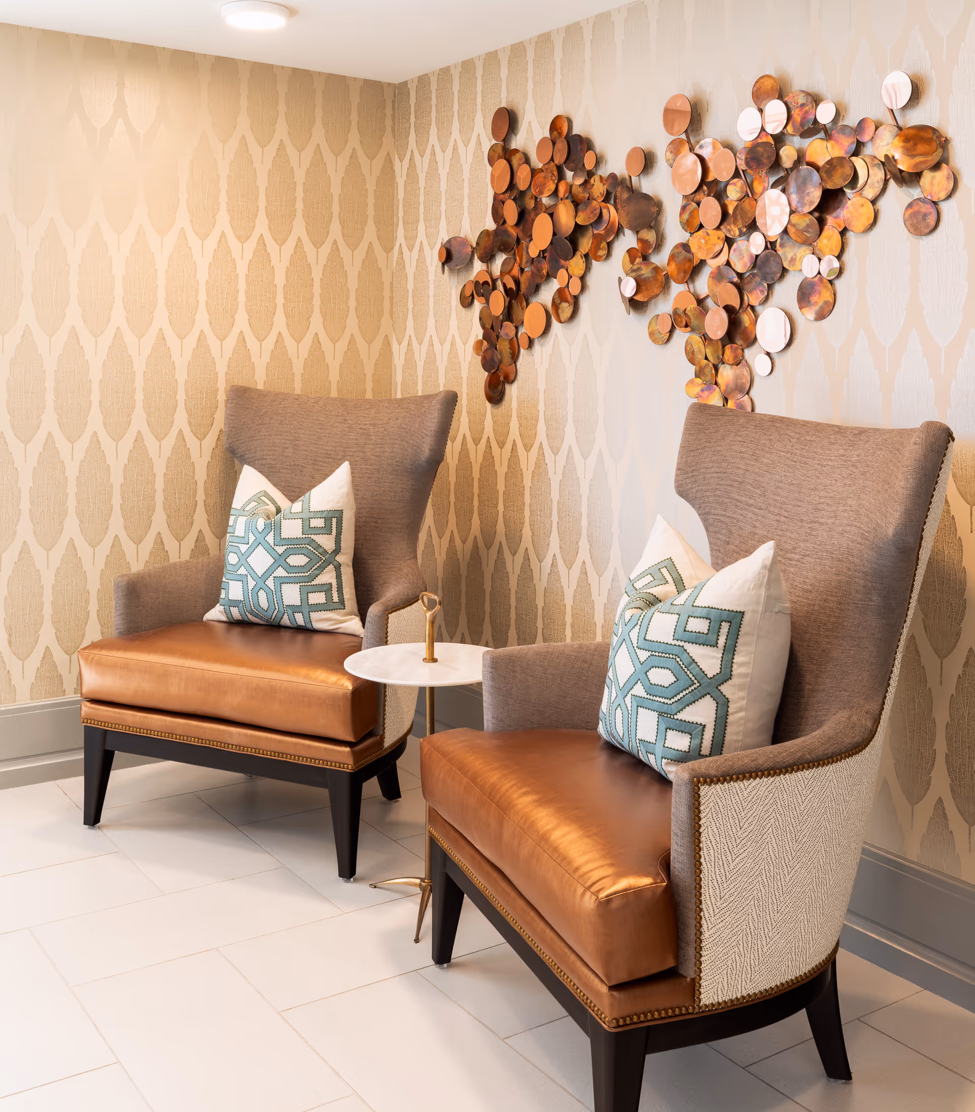 Two stylish armchairs with brown leather seats and patterned cushions are placed against a beige patterned wallpapered wall. Between the chairs is a small round white table with a gold handle. Above the chairs, there is a decorative wall art piece made of circular metallic elements in various shades of copper and bronze.