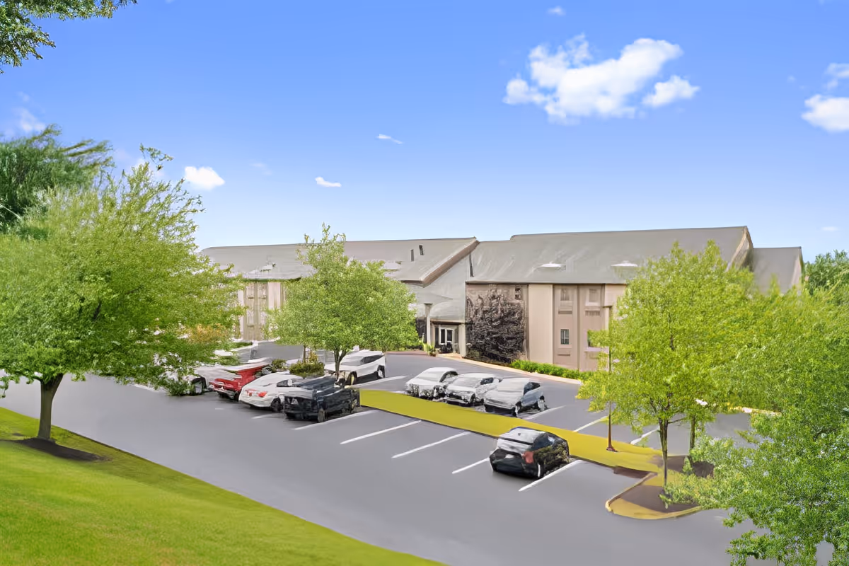 Exterior view of a senior living facility parking lot with several parked cars and green trees surrounding the area under a clear blue sky.