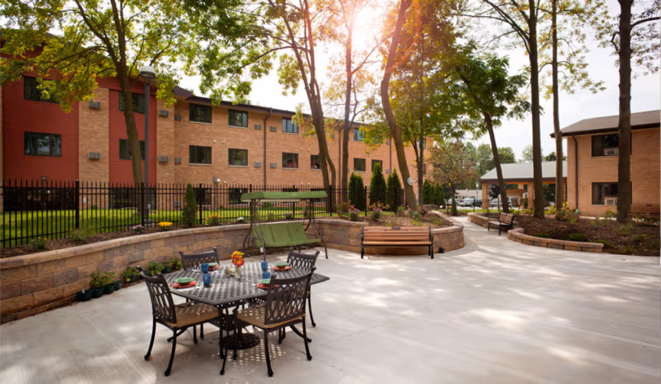 Outdoor patio area at Pine Haven Christian Communities Giddings Avenue Campus with a metal table and four chairs set with plates and cups. There are benches, a green cushioned swing, trees, and brick buildings in the background under a sunny sky.