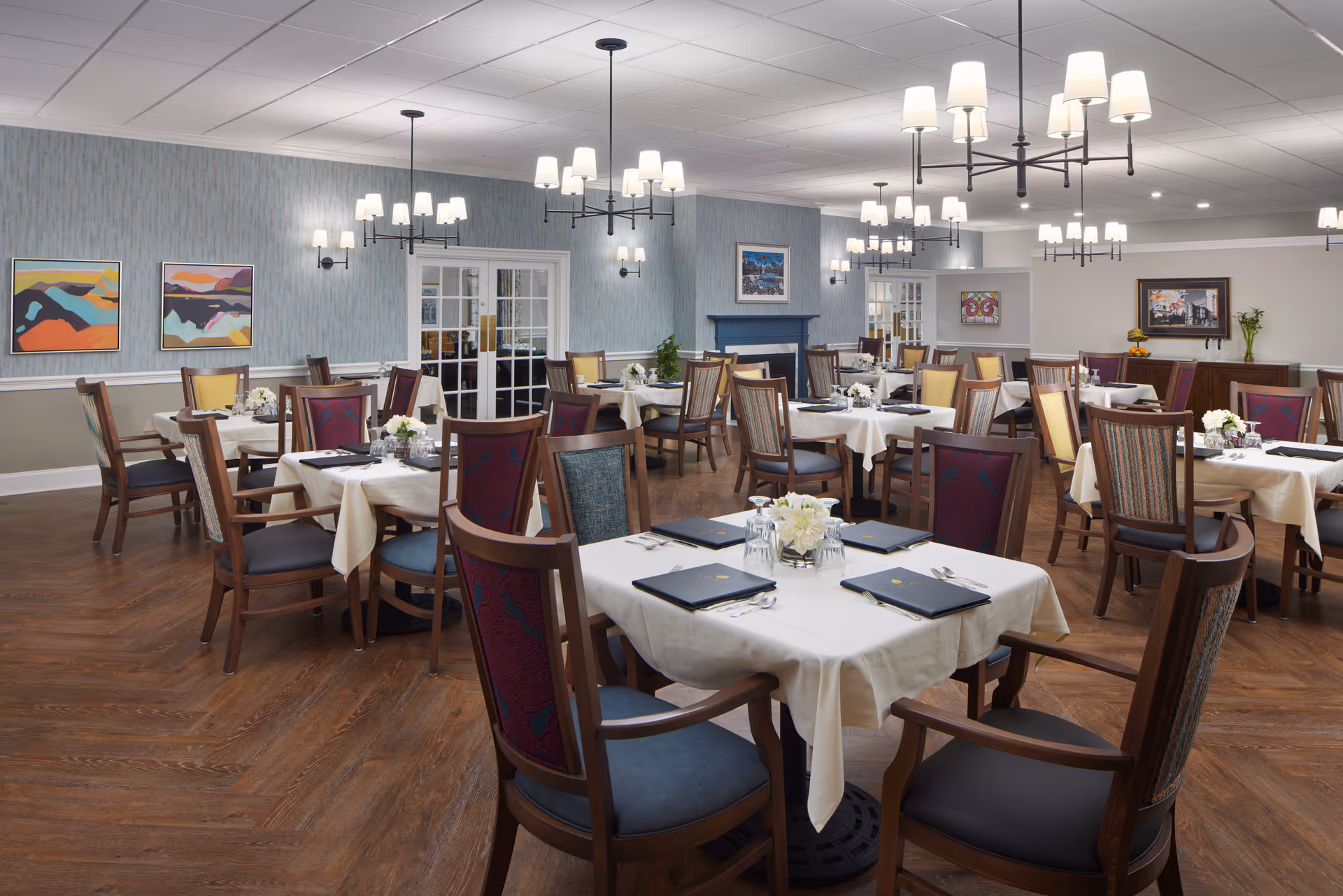 A spacious dining room with multiple tables covered in white tablecloths, each set with menus, glasses, and floral centerpieces. The room features wooden chairs with colorful upholstery, modern chandeliers, wall sconces, and framed artwork on the walls. The floor is wooden, and there are double glass doors in the background.