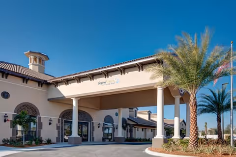 Front entrance of a Mediterranean-style memory care building with a covered porte-cochère, palm trees, and a clear blue sky.