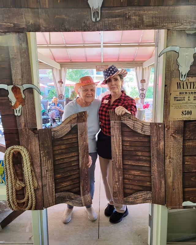 Two elderly women standing behind a wooden swinging saloon door prop, one wearing a pink cowboy hat and the other wearing a star-spangled cowboy hat and a red plaid shirt. The setting appears to be a covered outdoor patio with other people seated in the background and western-themed decorations including a rope and a wanted poster.