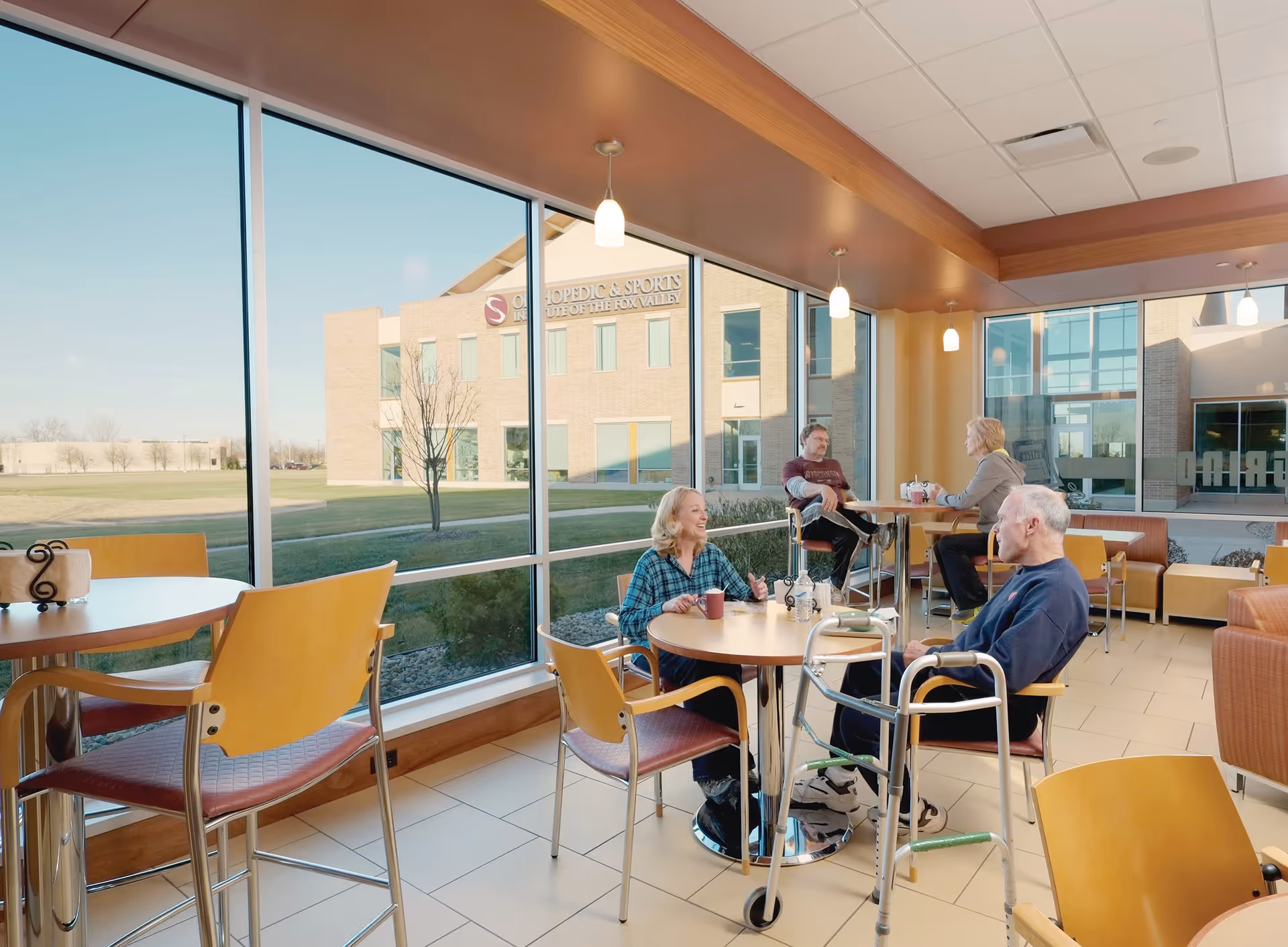 Bright common dining area with tables and chairs by large windows where several older adults chat and use walkers.