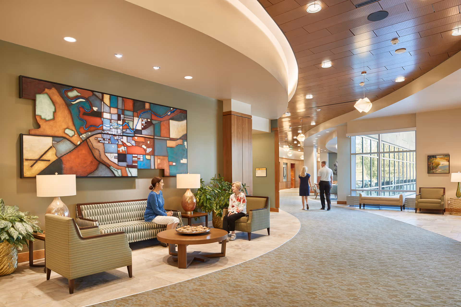 Bright curved lobby of a senior living facility with seating, large abstract wall art, plants, and residents conversing.