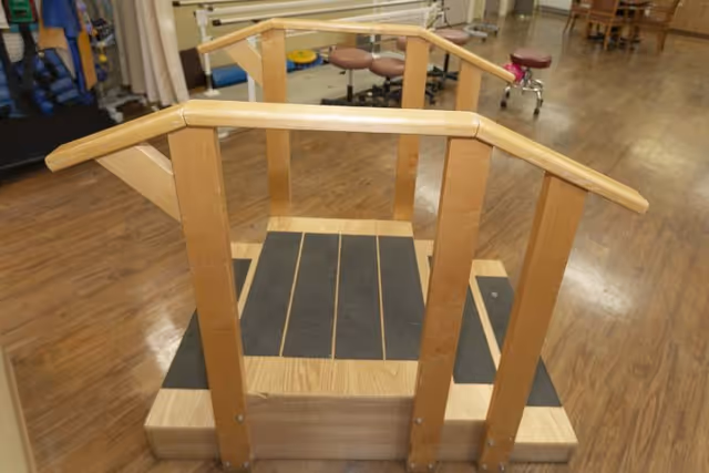 Wooden practice staircase with handrails on a hardwood floor in a therapy/rehab room.