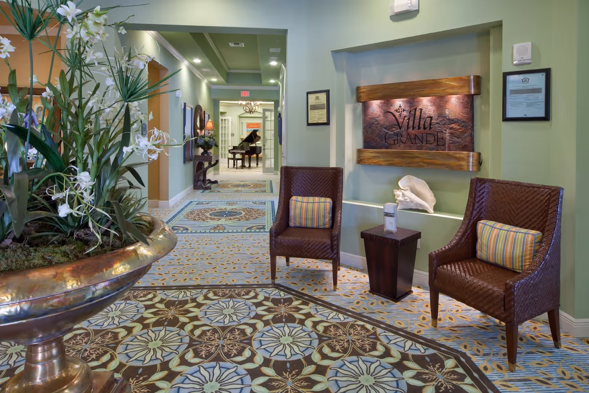 A welcoming interior seating area at Villa Grande on Saxon featuring two brown woven chairs with striped cushions, a small wooden side table, a decorative shell, and a wall plaque with the facility's name. The space has green walls, patterned carpet, and a large floral arrangement in a bronze planter. In the background, a hallway leads to a room with a grand piano.