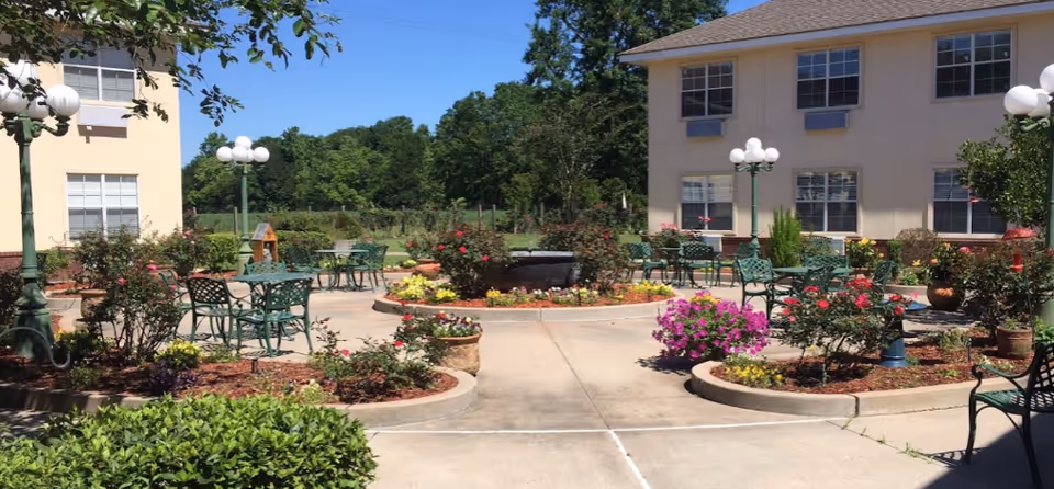 Outdoor courtyard area at Garden View Assisted Living & Memory Care featuring green metal tables and chairs, flower beds with colorful flowers, lamp posts with globe lights, and two beige buildings on either side under a clear blue sky.