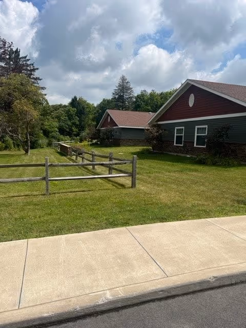 A grassy outdoor area with a wooden fence, a sidewalk, and two buildings with red roofs and dark green siding. Trees and a partly cloudy sky are visible in the background.
