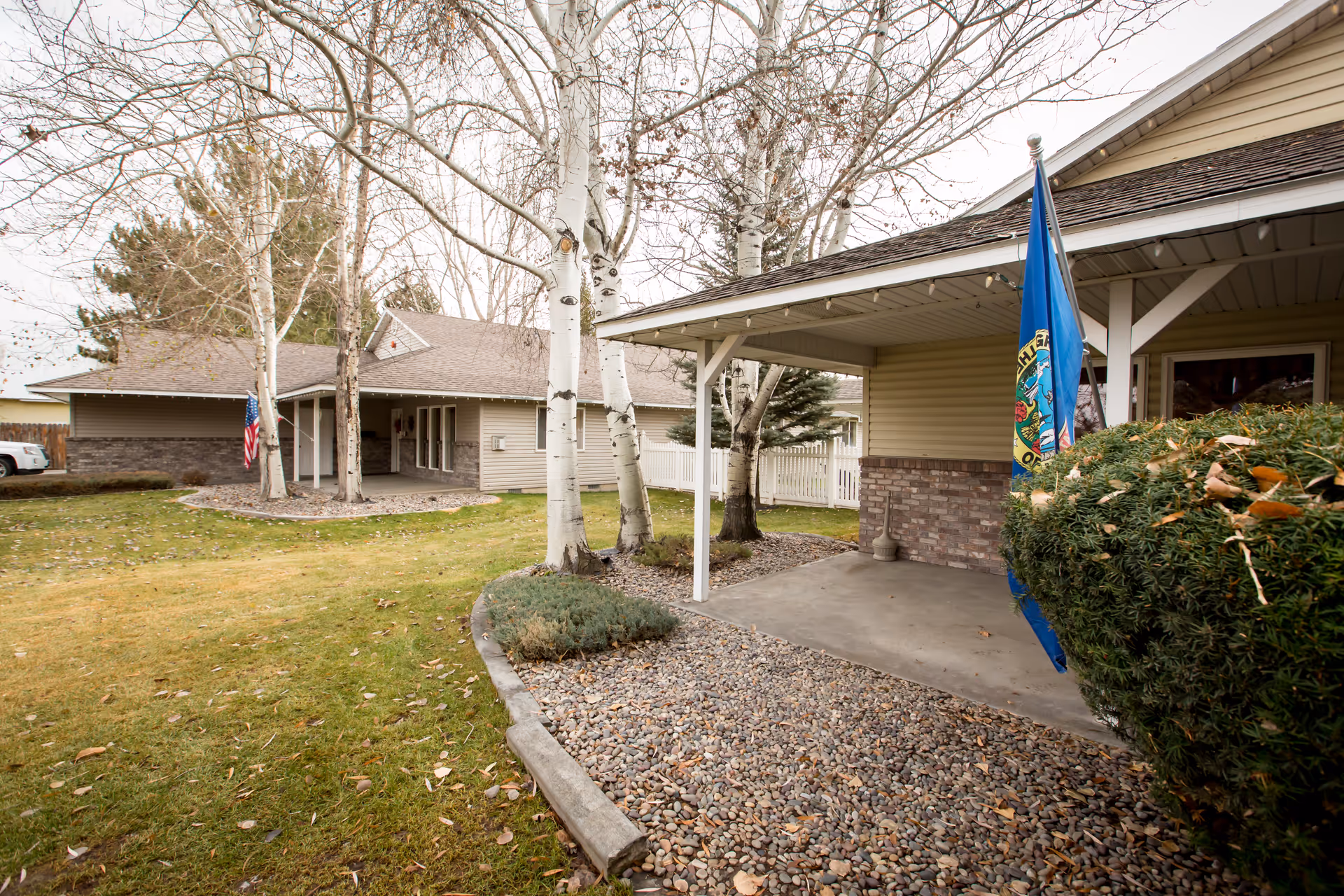 Exterior view of a single-story assisted living facility building with beige siding and a covered porch. There are leafless trees and a well-maintained lawn with some bushes and rocks in the landscaping. Two flags are visible, one American flag and one blue flag with a design. The sky is overcast.