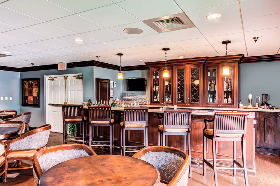 Interior view of a senior living facility bar area with wooden bar stools, a wooden bar counter, and a cabinet with glass doors displaying bottles and glassware. There are round tables with upholstered chairs in the foreground and pendant lights hanging from the ceiling.