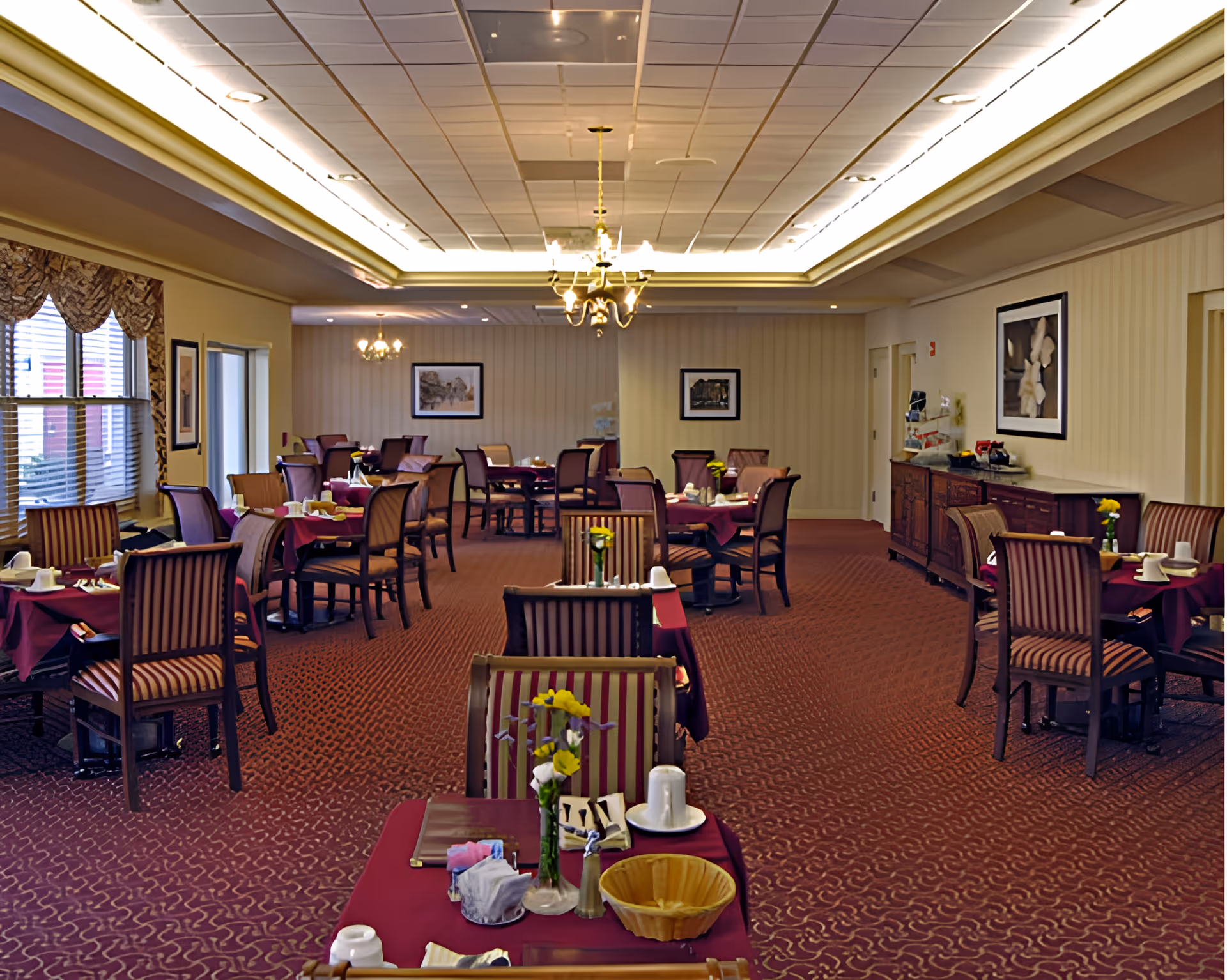 A dining room with multiple tables covered in burgundy tablecloths, each set with cups, plates, and small flower vases. The room has striped upholstered chairs, patterned carpet, framed artwork on the walls, and chandeliers hanging from a recessed ceiling with soft lighting.