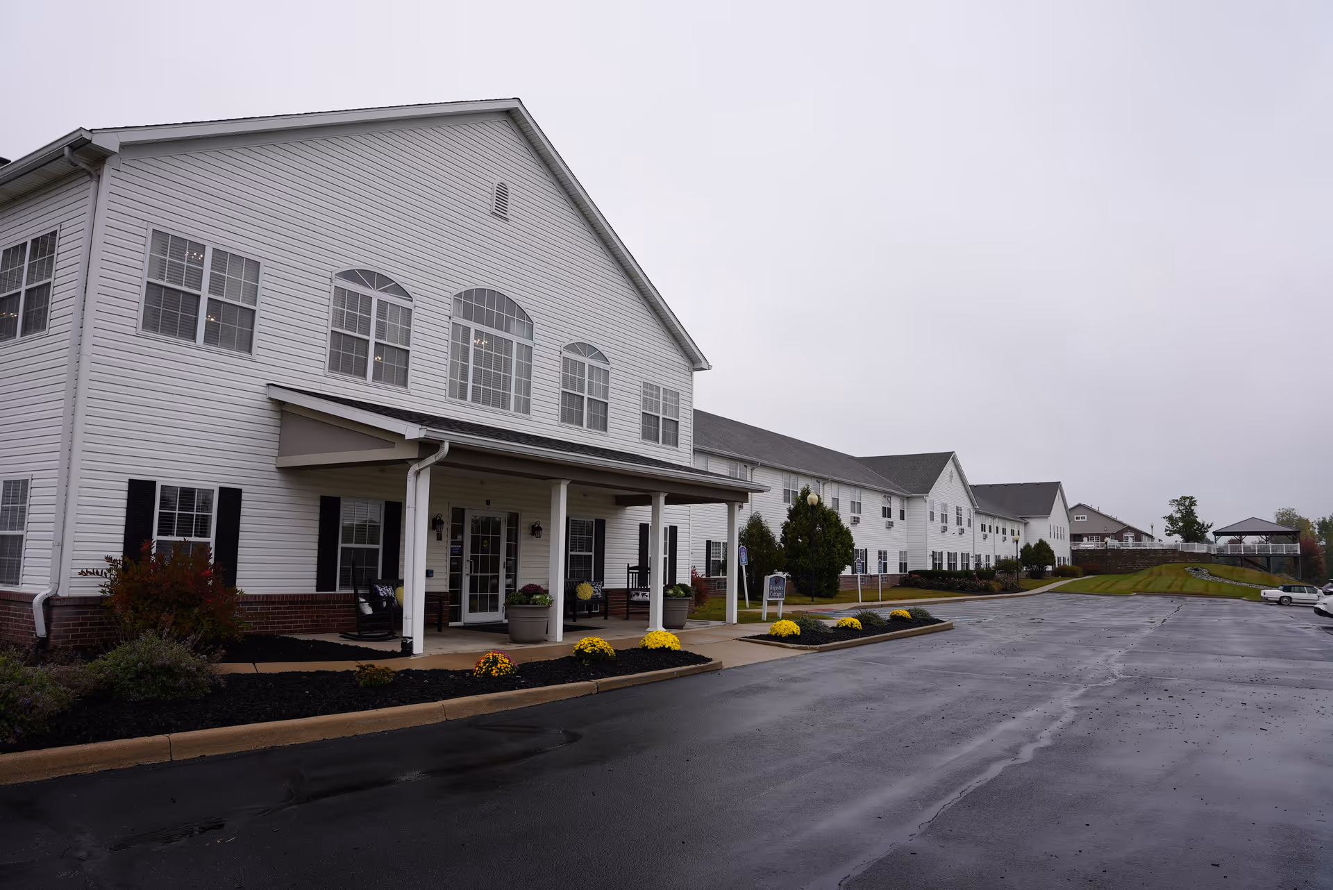 Exterior view of a large white two-story assisted living facility building with multiple windows and a covered entrance. The building is surrounded by a paved parking lot and landscaped areas with flowers and shrubs under an overcast sky.