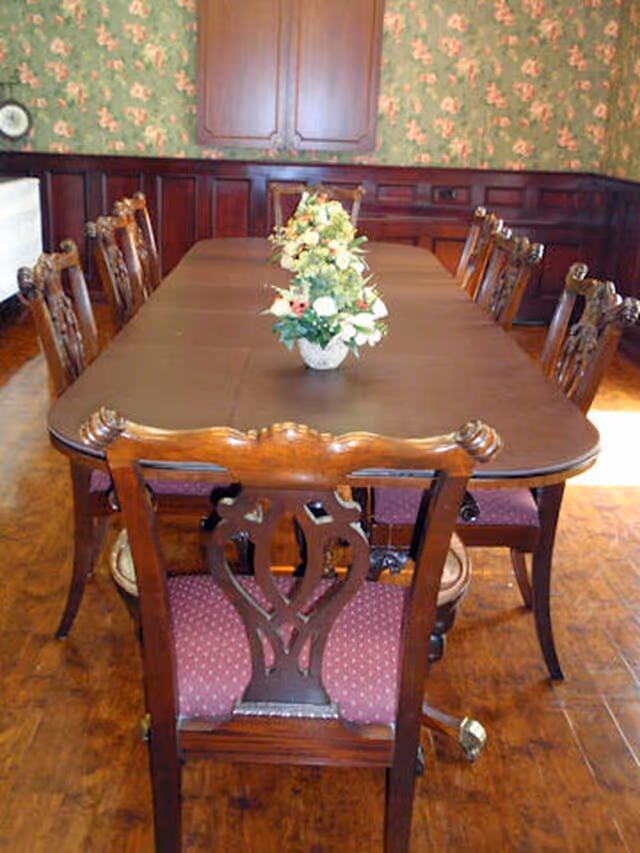 A traditional dining room with a long wooden table surrounded by ornate wooden chairs with red cushioned seats. A floral centerpiece is placed in the middle of the table. The room has floral wallpaper and wooden paneling on the walls.