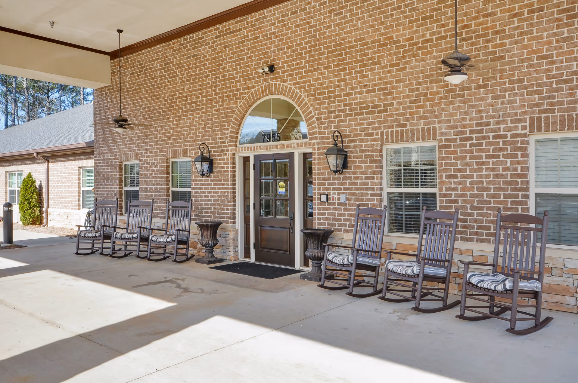 Covered outdoor patio area at Oaks at Shiloh Point with a row of wooden rocking chairs with striped cushions lined up against a brick wall. The entrance door has an arched window above it and is flanked by two large wall-mounted lantern lights. Ceiling fans hang from the covered patio ceiling.