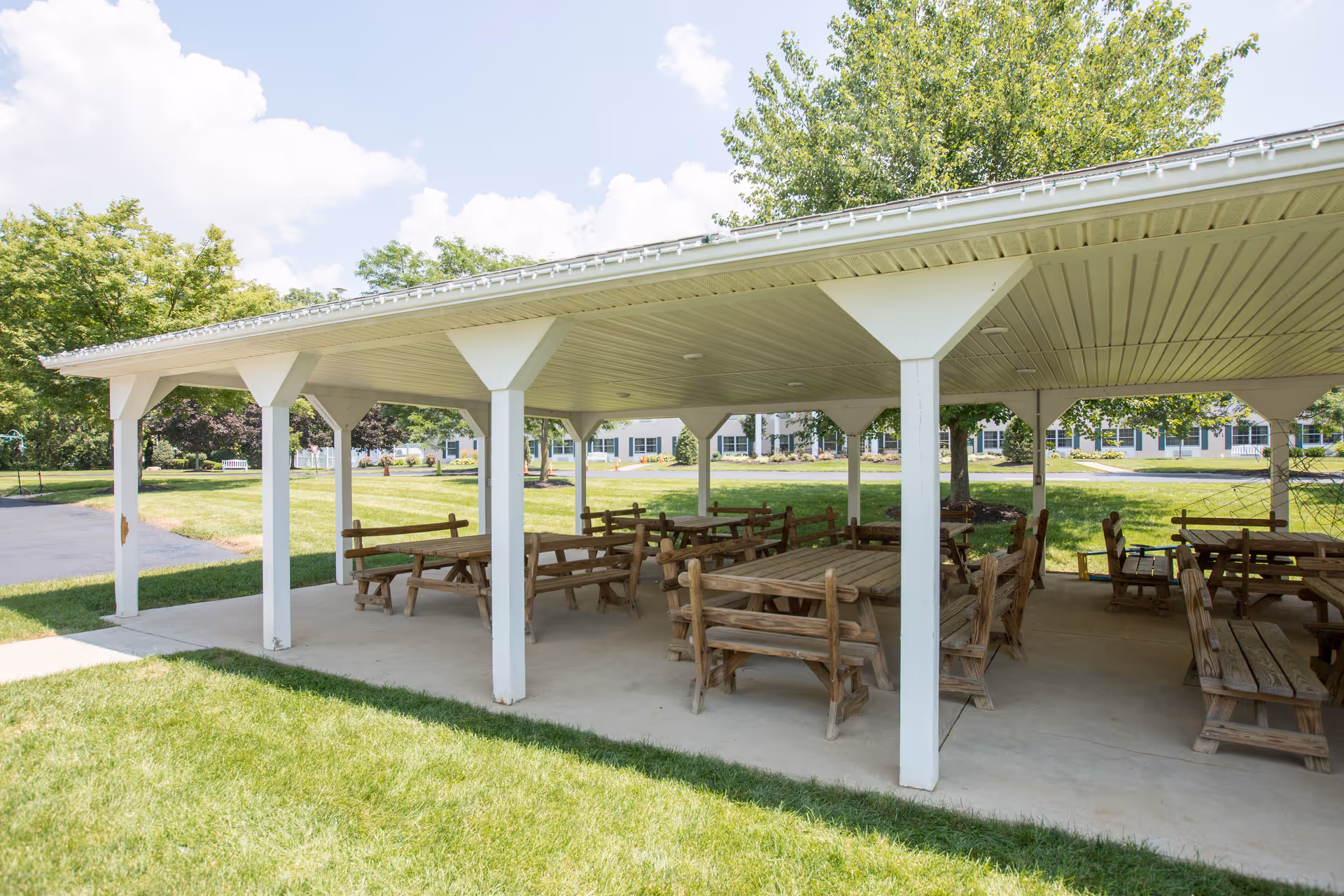 Outdoor covered pavilion with multiple wooden picnic tables and benches on a concrete floor, surrounded by green grass and trees, with a building visible in the background under a partly cloudy sky.