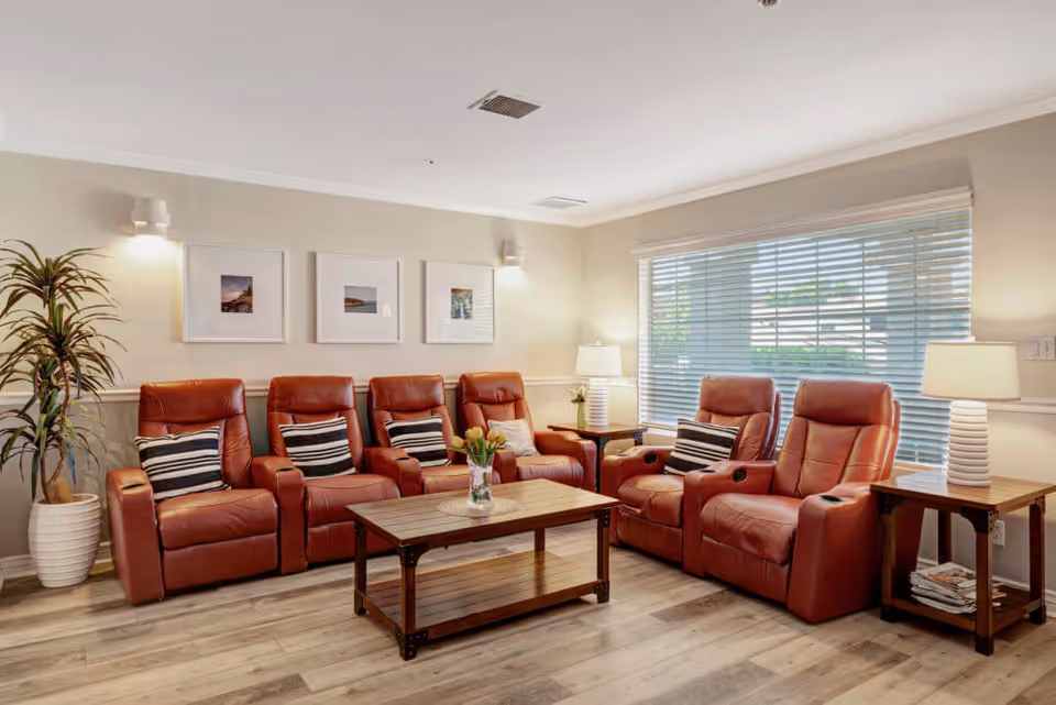 A cozy living room with six red leather recliner chairs arranged in a row, each with a striped pillow. There is a wooden coffee table with a vase of flowers in the center, two wooden side tables with white lamps on either side of the chairs, a large window with blinds letting in natural light, three framed pictures on the wall, and a tall potted plant in the corner.