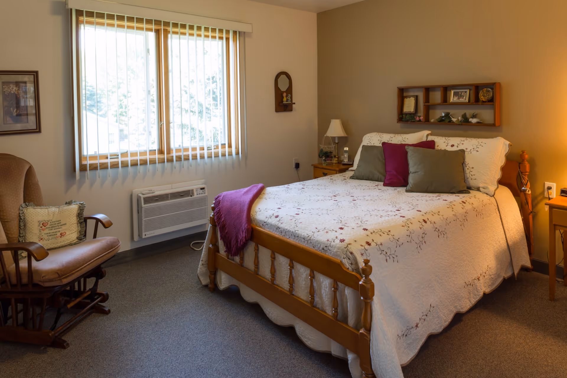 A cozy bedroom with a wooden bed frame, a white floral quilt, and several pillows in green and purple. There is a wooden rocking chair with a cushion to the left, a window with vertical blinds, and a small air conditioning unit below the window. On the right side of the bed, there is a nightstand with a lamp and a phone. A small wooden shelf with decorative items is mounted on the wall above the bed.